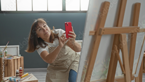 Foto Middleaged woman painter in apron holds smartphone and taps screen in studio near easel and paint jars; focused creativity