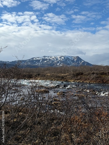 Blaskogabygg, Iceland - May 08, 2026: Snow-capped mountain rises above a river with rocky banks and sparse vegetation under a cloudy sky in Icelandic landscape