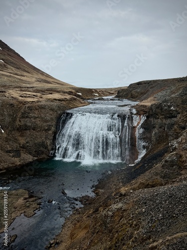 Kjosarhreppur, Iceland - May 08, 2026: Waterfall cascading down rocky cliffs into a river surrounded by barren landscape under a cloudy sky in Iceland