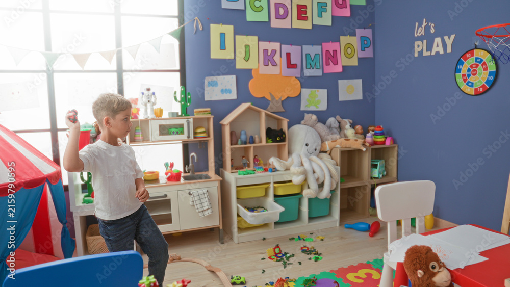 Fototapeta premium Boy throws a small red ball with his right hand inside a colorful children's playroom building scattered with blocks, tent and stuffed toys; playful joy.