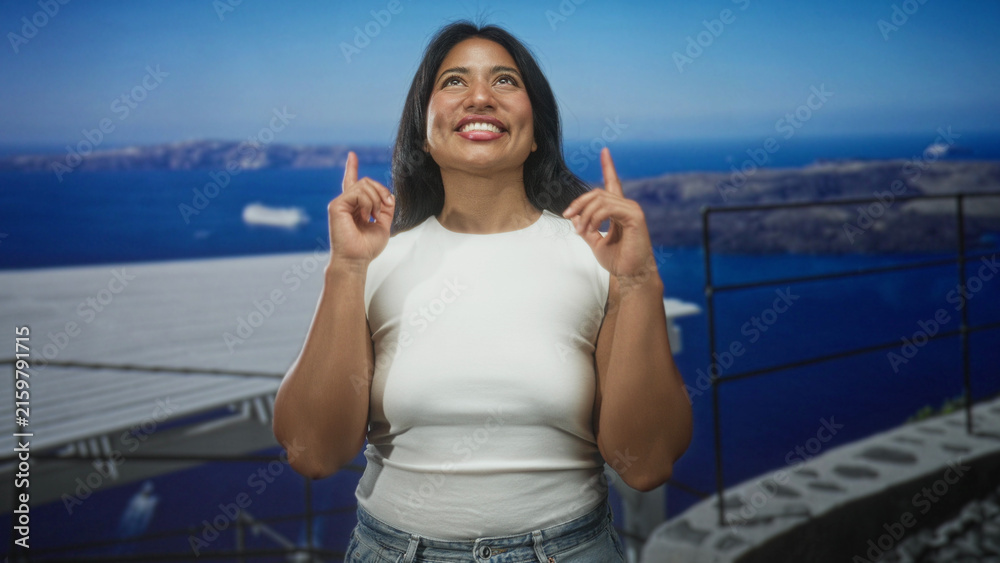 Fototapeta premium Woman pointing index fingers upward on seaside balcony in white sleeveless top and jeans; joy optimism.