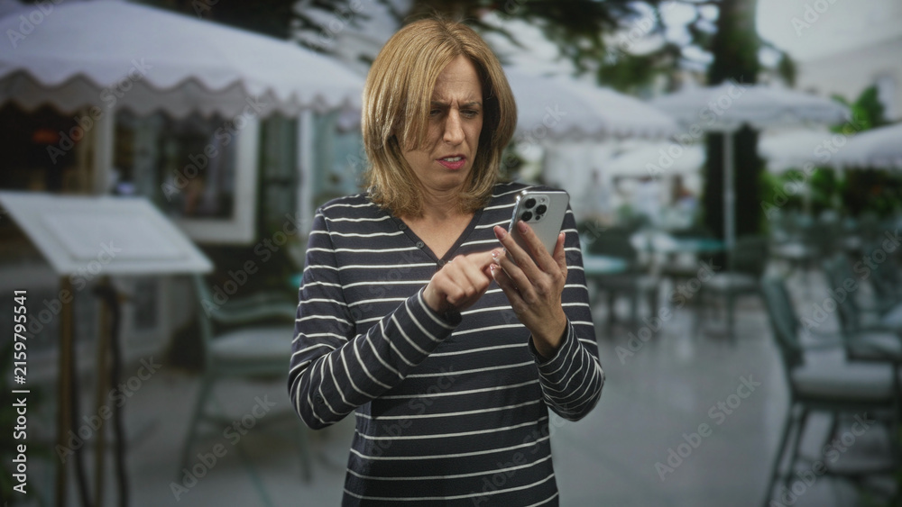 Fototapeta premium Woman tapping smartphone with visible hands and furrowed expression at a street cafe terrace, holding phone while scrolling through messages; everyday tech frustration.