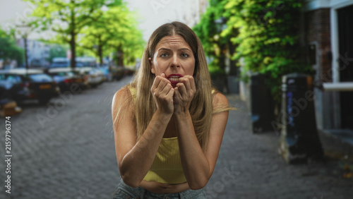 Konstfotografi Woman with hands clenched in a pleading gesture on a cobblestone street, wearing yellow crop top and jeans; anxiety vulnerability