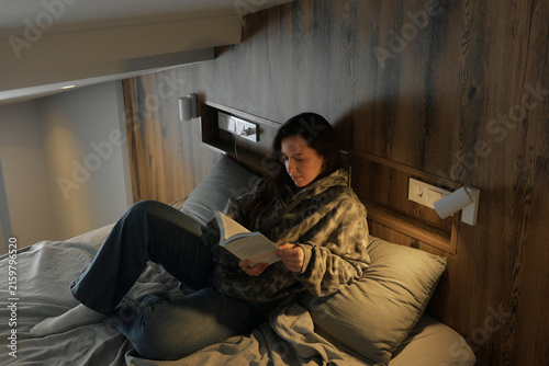 Woman reading a book while sitting on a bed in a cozy room during the evening hours