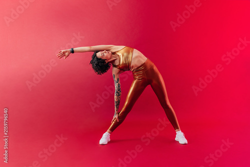 Yong woman in golden sportswear stretching her body over crimson backrground in studio