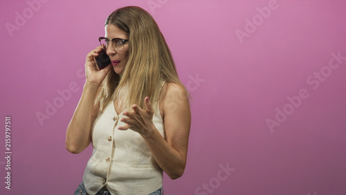 Photography Woman holds smartphone to ear, smiles and gestures with hand while wearing glasses and a buttoned vest against a pink studio backdrop; joyful connection