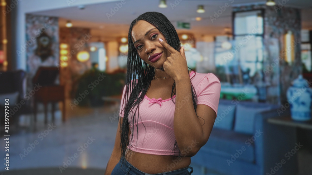 Fototapeta premium Black woman pointing finger to cheek and eye, wearing pink crop top in a building lobby; confidence empowerment poise.