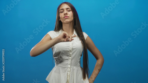 Canvastavla Young woman in white dress points finger with raised hand, lips pursed and long hair visible, studio; confidence assertiveness