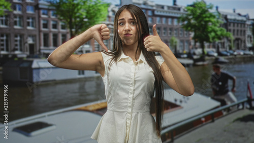 Photography Teenage girl wearing white sleeveless dress shows thumbs up and thumbs down with visible thumb gesture by canal boat and building; ambivalence