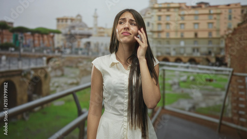 Foto Young woman on phone wiping tears with hand to face in white dress on building balcony; sadness solitude