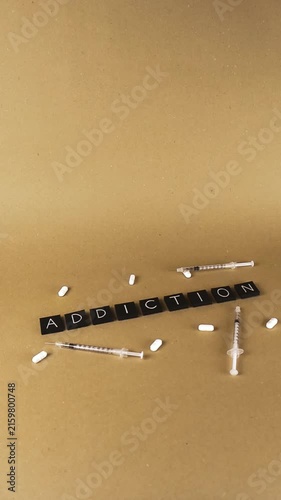 Conceptual shot showing the word addiction spelled with black tiles, surrounded by white pills and syringes, representing substance abuse, dependency, and the dangers of drug misuse