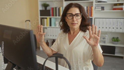 Quadro su tela Middle age woman hands raised palms out at computer desk in office with keyboard and monitor visible; defensive concern