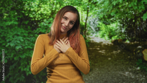 Young redhead woman wearing mustard sweater with hands on chest smiling on a forest path lined with green foliage; gratitude reflection.