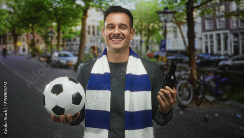 Man holding soccer ball and beer bottle, wearing blue and white scarf, looking hesitant on street; ambivalence support team spirit.