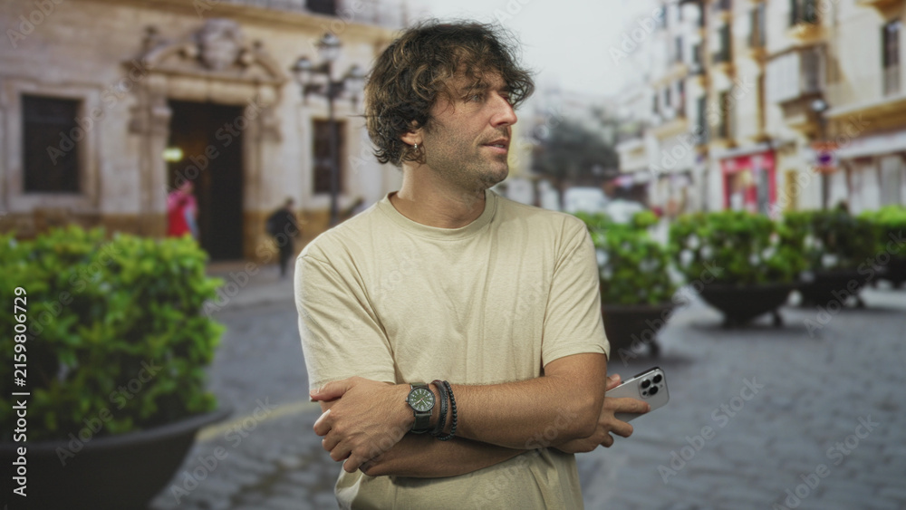 Fototapeta premium Man with smartphone and arms crossed on a cobblestone street by planters and a historic building; contemplation travel.