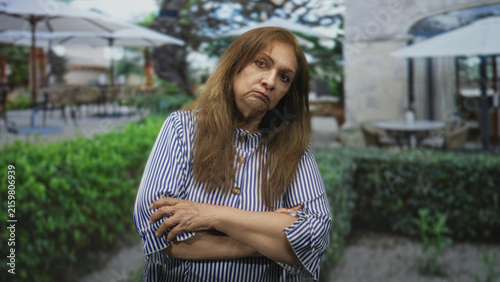 Cuadro en lienzo Woman with crossed arms and stern expression wearing striped shirt by patio umbrellas, tables and hedge at a building courtyard; disapproval solitude