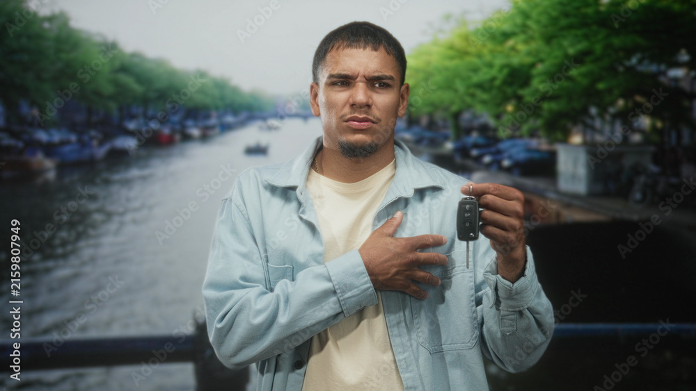 Fototapeta premium Young hispanic man holding a car keyfob, hand pressed to chest on a street canal bridge; concern uncertainty about a lost or broken car key.