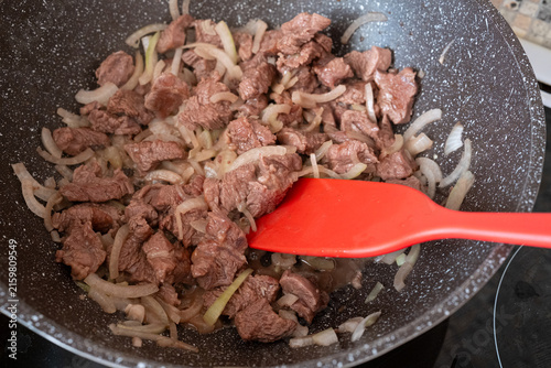 beef pieces stewing with sliced onion in deep frying pan. spatula mixing beef during cooking of tasty homemade meal. concept of beef dish and family dinner preparation at home kitchen