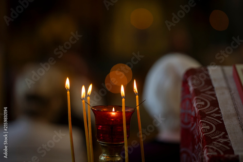 Lit beeswax candles surrounding a red glass vigil lamp in an ornate metal candle stand inside a church, creating a warm and contemplative atmosphere for prayer and worship. 