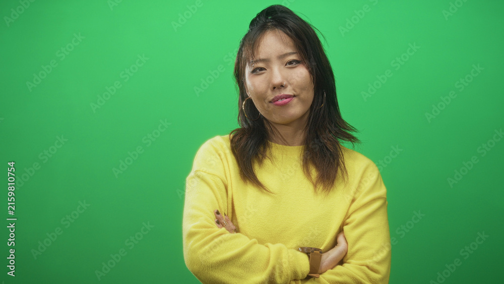 Fototapeta premium Woman with arms crossed wearing yellow sweater, visible face and hoop earrings on green screen studio; confidence poise.