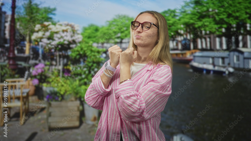 Fototapeta premium Blonde woman with clenched fists near chin on street by canal, wearing glasses and a pink striped shirt; playful confidence.