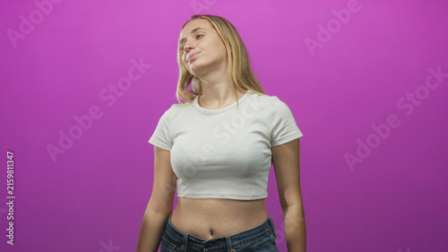 фотографія Woman tilting head with puckered lips showing exposed stomach and navel in studio against magenta backdrop wearing white crop top and jeans; confident attitude