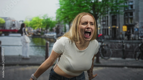Woman shouting with open mouth and visible midriff in a cropped white tshirt on a city street by a canal and bridge; frustration release.