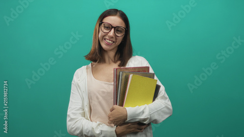 Obraz na płótnie Woman smiling while holding a stack of colorful notebooks and textbooks, hands clasped across chest wearing round glasses in studio; cheerful focused learning