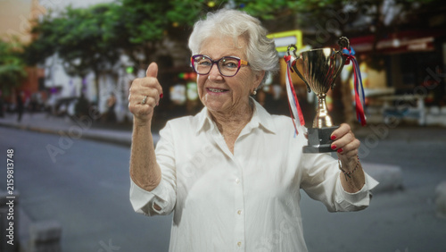 Konstfotografi Woman holding a trophy and giving thumbs up on a busy street, visible hand and smiling face with glasses; pride joy