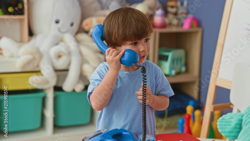 Quadro su tela Boy holding a toy burger with both hands while reaching toward a pretend kitchen shelf and stuffed octopus in a colorful playroom inside a building; playful childhood moment