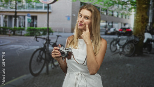 Fotografía Woman in white dress holding vintage camera, index finger touching cheek, posing on street by bicycles and parked car; travel memory playful