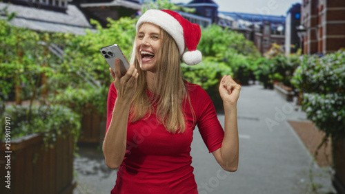 Foto Blonde woman speaking into smartphone, holding phone to mouth and smiling with a raised fist on street lined with planters and brick buildings; joyful holiday celebration