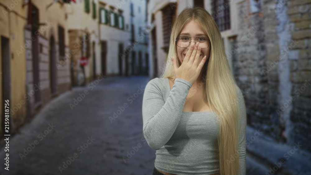 Fototapeta premium Young blonde woman with glasses covers mouth with hand on narrow cobblestone street; surprise reaction.