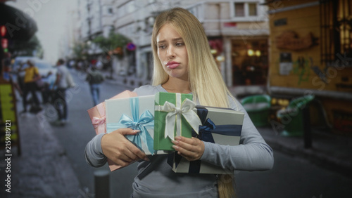 Quadro su tela Young blonde woman holding several wrapped gift boxes tightly against her hands, pouting lips on a busy city street; frustration