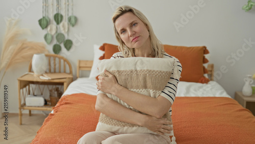 Woman hugging pillow on bed inside a building, hands clasped over cushion and orange throw, seated in gentle posture; quiet contemplation solitude.