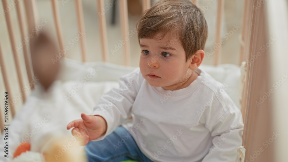 Fototapeta premium Boy reaches with bare hand toward hanging plush mobile above crib in a residential building nursery, face intent and focused on the toy; curiosity.