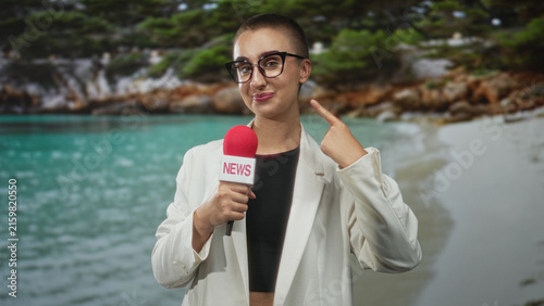 Cuadro en lienzo Woman holding red microphone labeled news points index finger to cheek while smiling at camera on a sandy beach cove wearing glasses white blazer black crop top; broadcast confidence