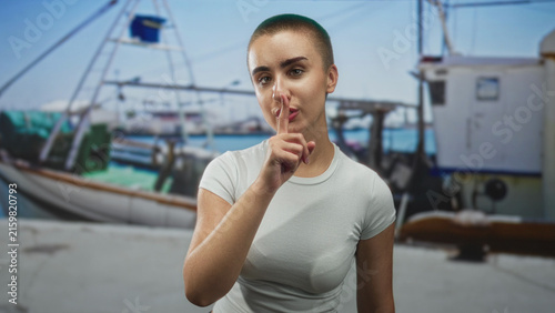 Fotografía Woman with short hair places finger to lips in a marina harbor by a fishing boat and dock; quiet secrecy