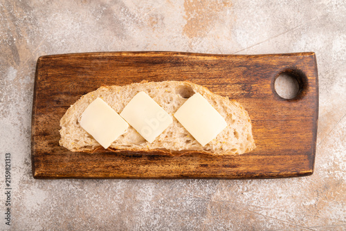 Slice of homemade bread with butter on cutting board on brown concrete, close up, top view.