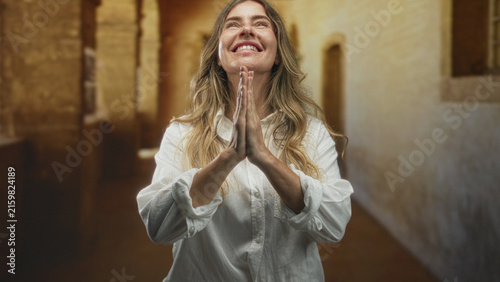 Konstfotografi Woman with clasped hands pressed together in prayer, looking upward inside a stone building with white shirt and golden sunlight; faith hope