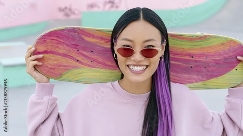 Cheerful young woman with colorful skateboard held over shoulders in a pastel skatepark for portrait background and social media banner use