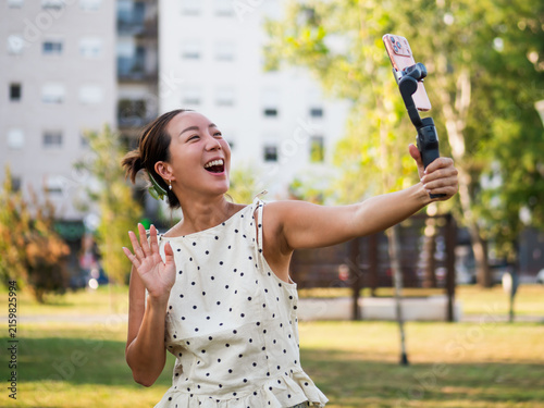 Happy mid adult woman waving at the camera while recording a cheerful selfie video with a smartphone stabilizer outdoors.