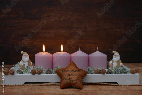 Einen schönen zweiten Advent. Rustikales Gesteck dekoriert mit Weihnachtsmann Figuren, Stern und Tannenzweigen vor einem rustikalem Holzhintergrund mit Platz für Text.