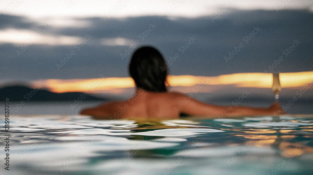 Fototapeta premium Woman relaxing alone in an infinity pool overlooking the ocean at sunset, enjoying a luxury travel experience with a drink
