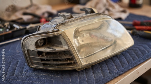 Used dusty automotive headlight assembly with a faded clear lens resting on a blue quilted pad on a workbench