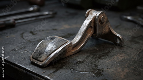 Used dirty metallic automotive foot pedal assembly resting on a dark greasy workbench
