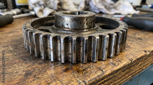 Used oily metal engine timing sprocket resting on a scratched wooden workbench in a mechanic garage