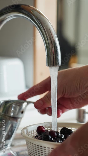 Close up of hands washing dark berries in white colander. Hands move colander to rinse contents with water dripping. Background shows typical kitchen environment