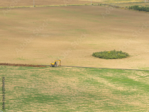 Yellow excavator digging a trench for pipeline installation in vast rural agricultural field under bright daylight, highlighting infrastructure development and earthmoving work.