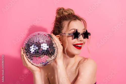 Photography Young woman with a disco ball and star sunglass poses on a vibrant pink backgrou